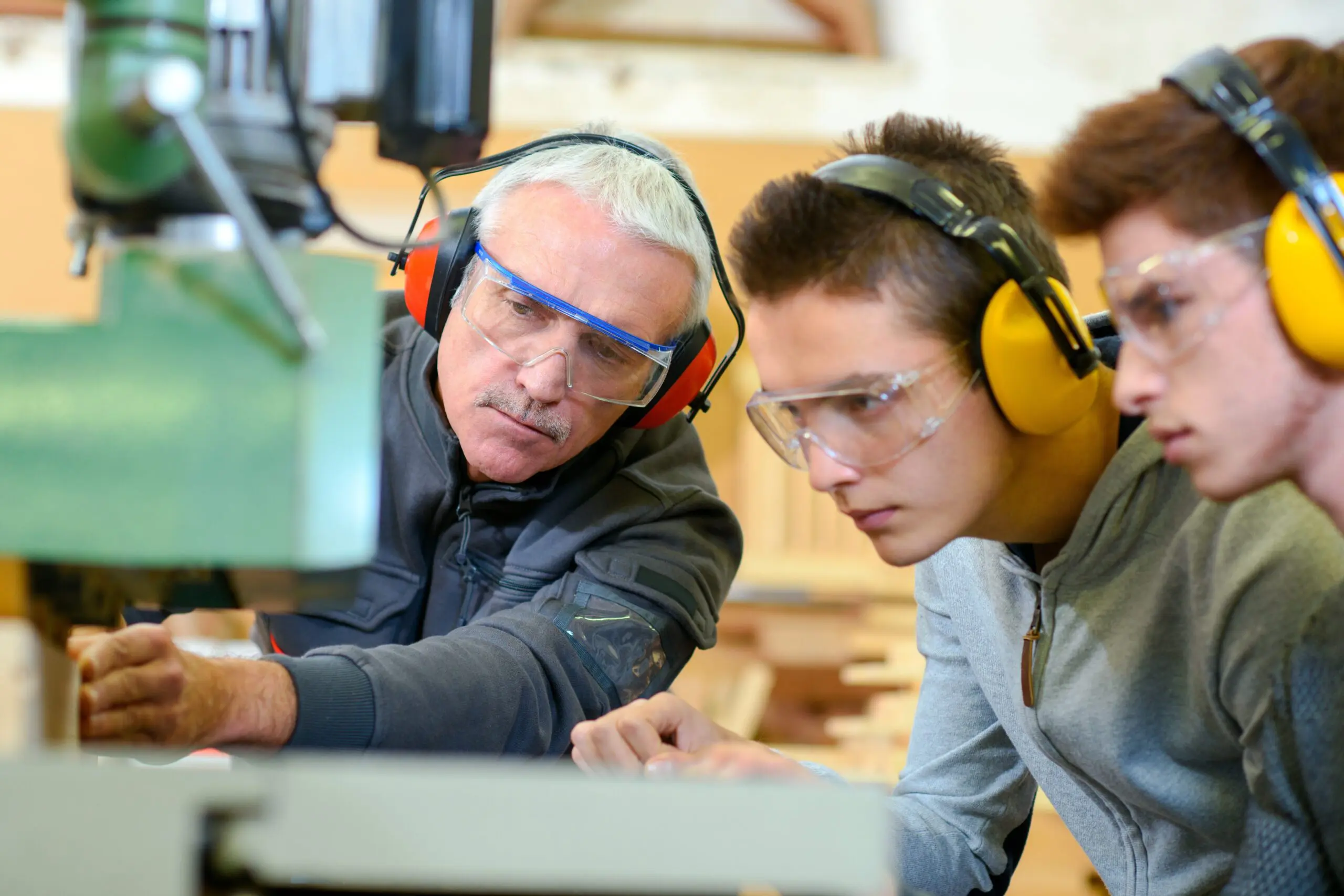 An older man teaches two young men woodworking; all wear safety glasses and ear protection.