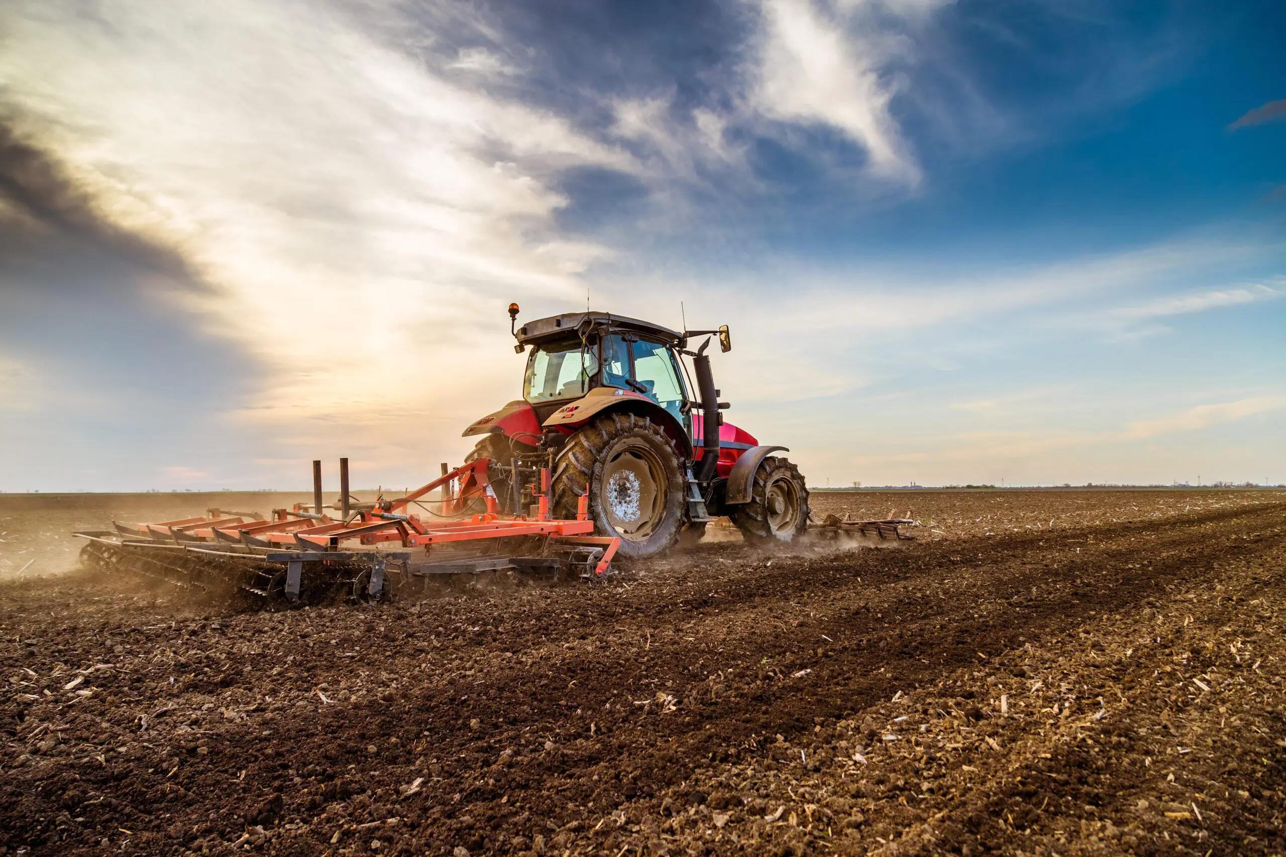 A red tractor plowing a large field under a cloudy sky at sunset.