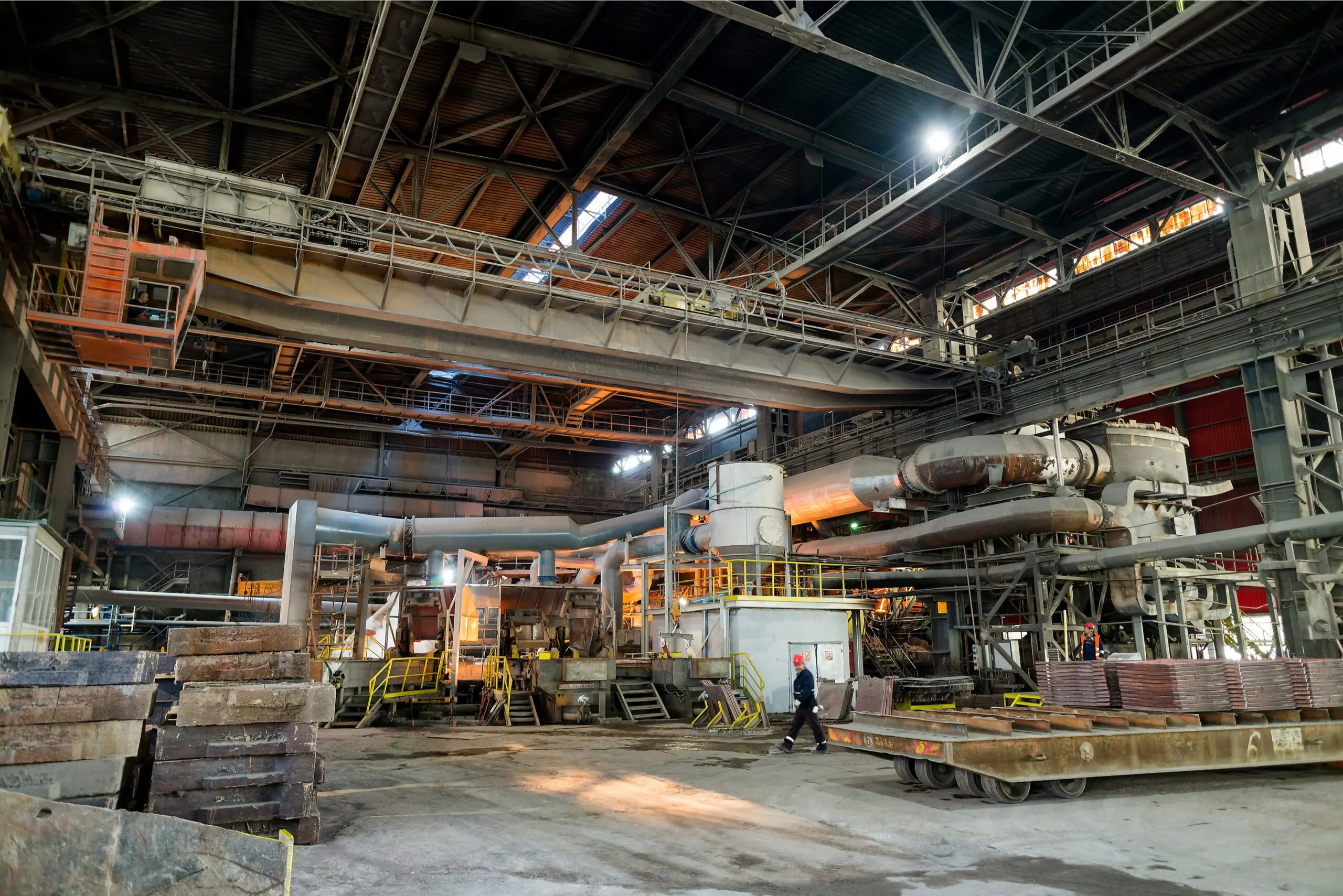 Interior of a large industrial factory with machinery, metal platforms, and a worker walking on the floor.