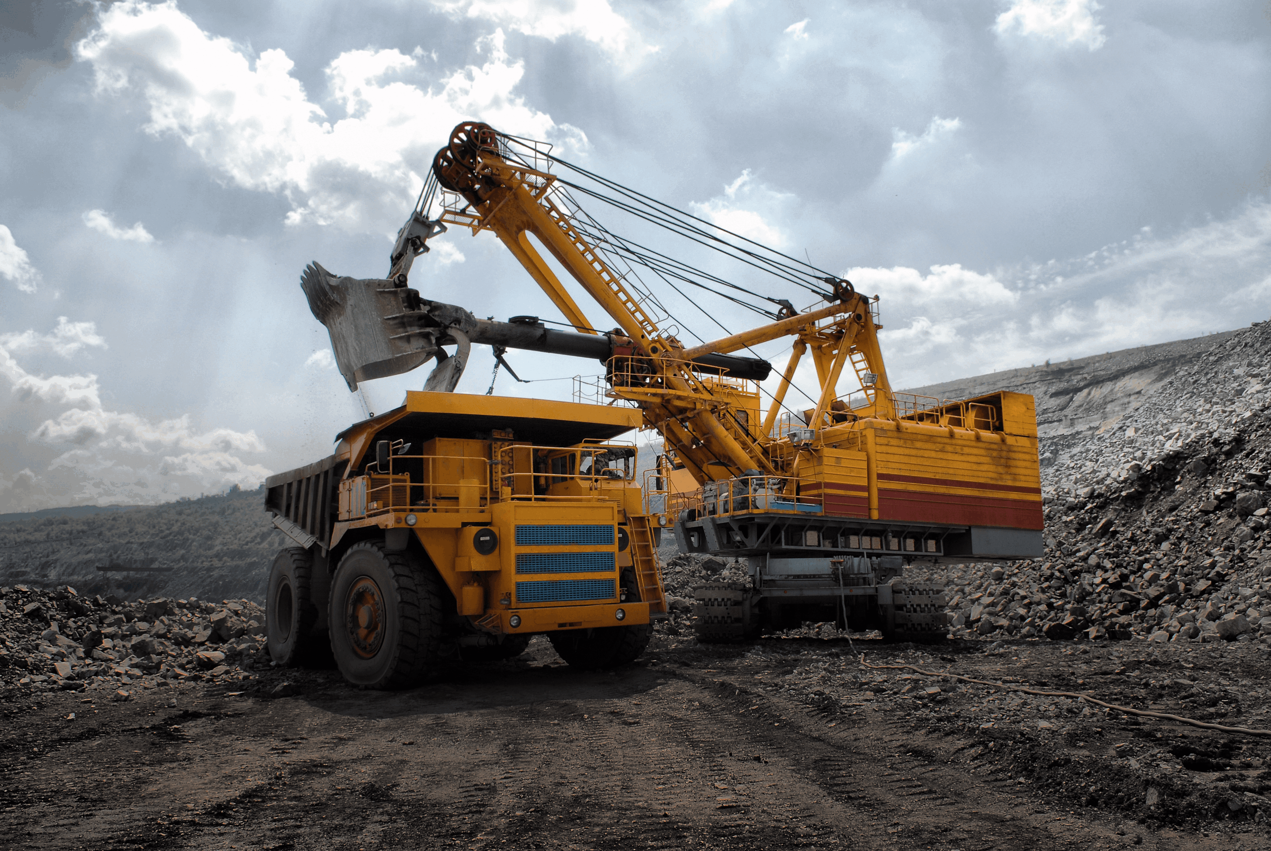 A large excavator loads rocks into a dump truck at an open-pit mining site under a cloudy sky.