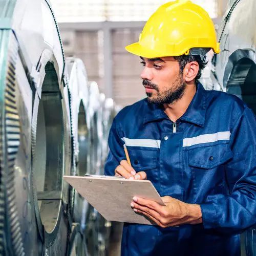 Man in safety helmet and coveralls inspects machinery and takes notes on a clipboard in an industrial setting.