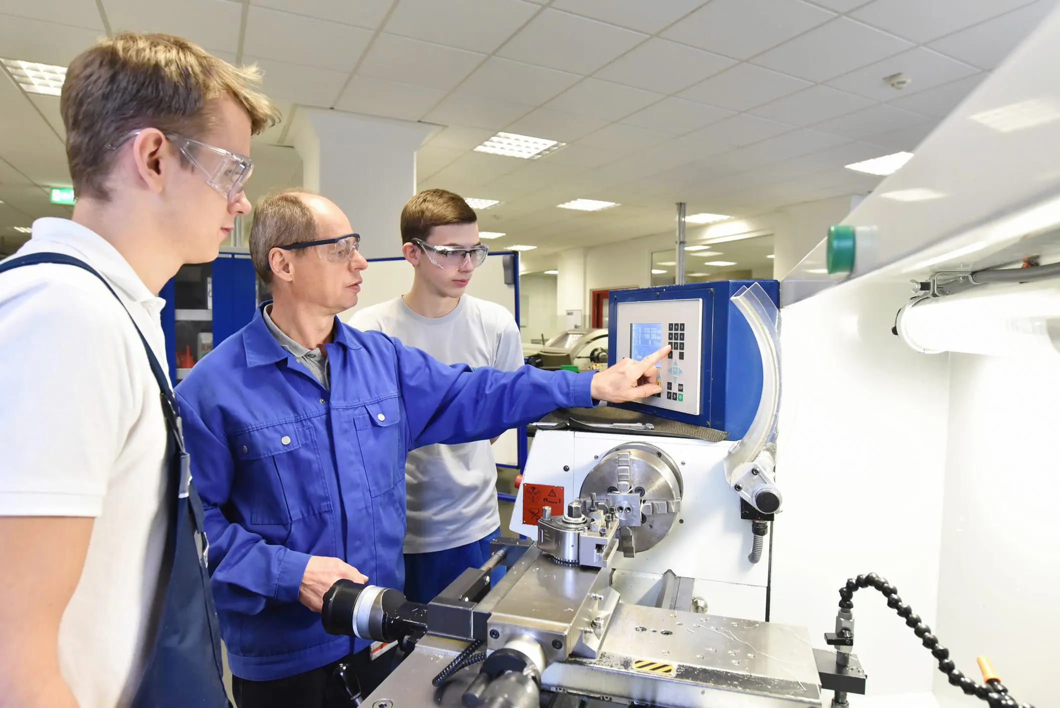 Three men in safety glasses operate and view a control panel on a machine in a bright workshop.