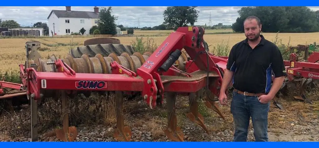 A man stands by a red farm cultivator in a field with a white house and trees in the background.