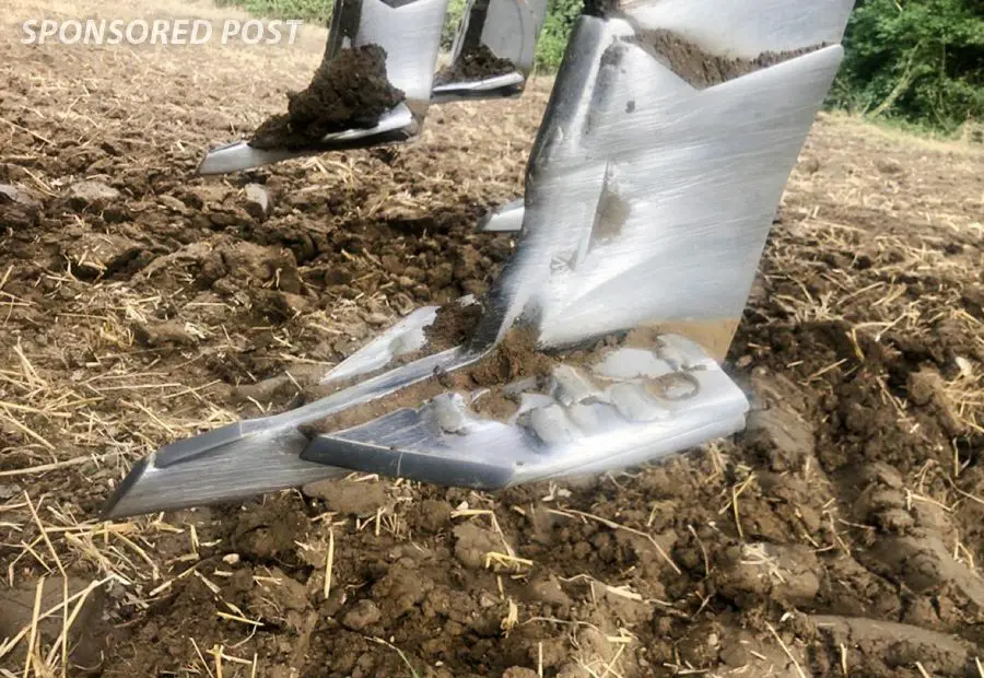 Close-up of metal agricultural plow blades with soil and straw on a freshly tilled field.