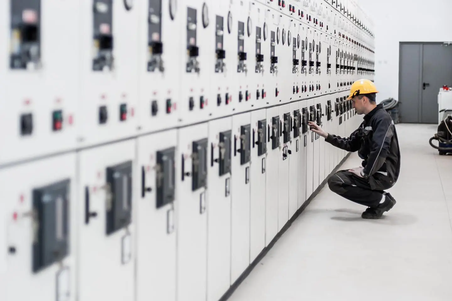 Worker in a hard hat operates control panels in an industrial facility, kneeling near electrical cabinets.