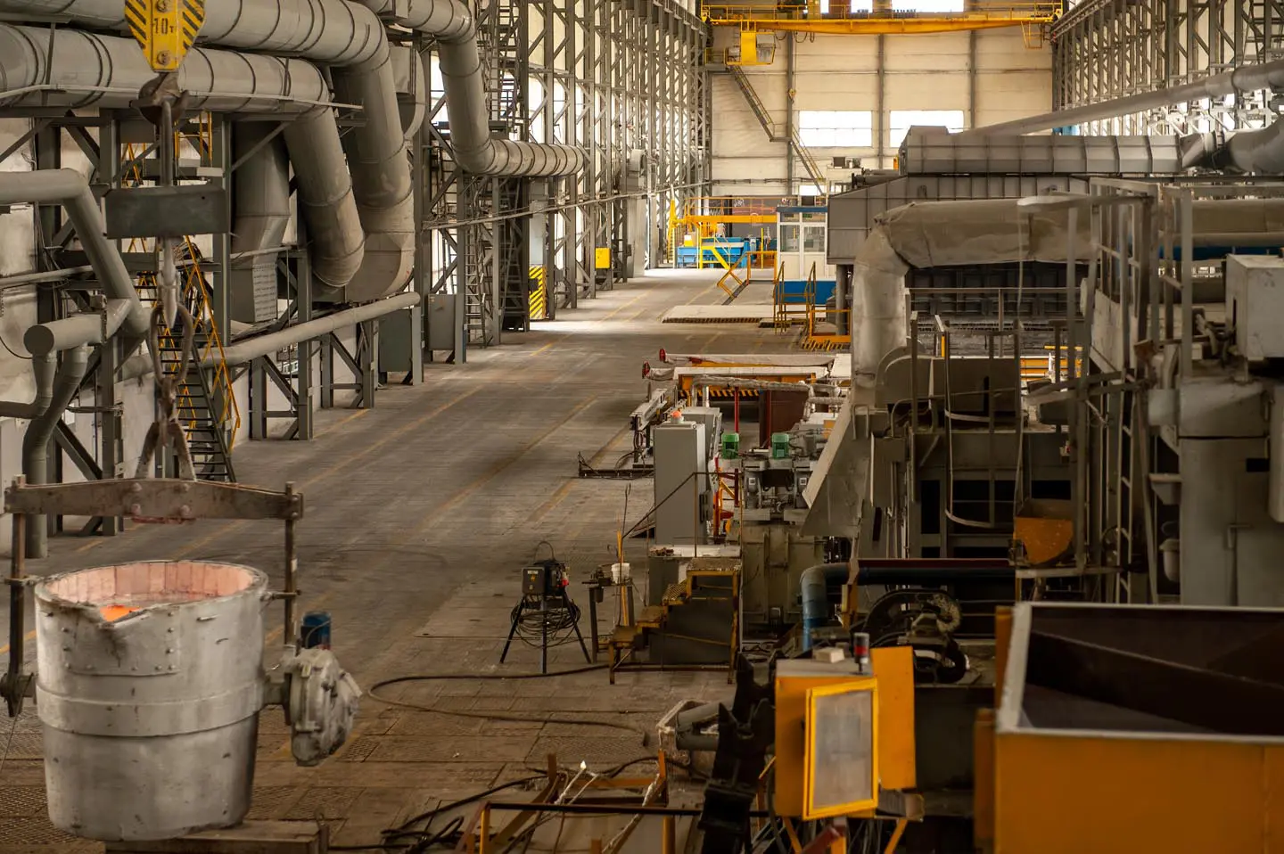 Large industrial factory interior with machinery, metal pipes, and a container of molten material in the foreground.