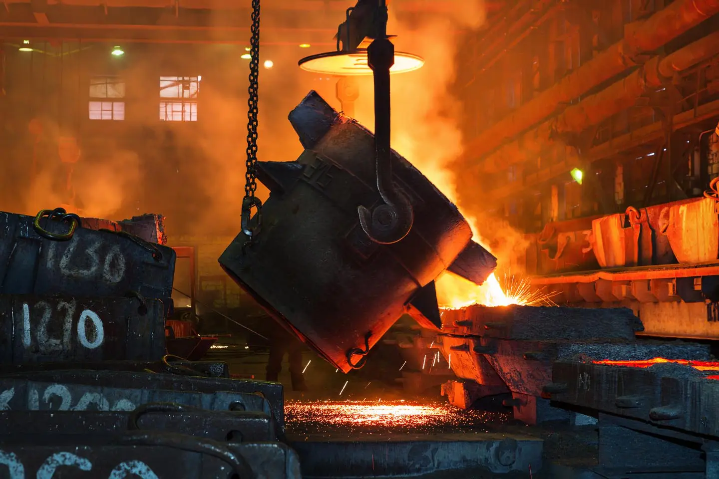 Molten metal is poured from a large ladle in a glowing, smoky industrial foundry.