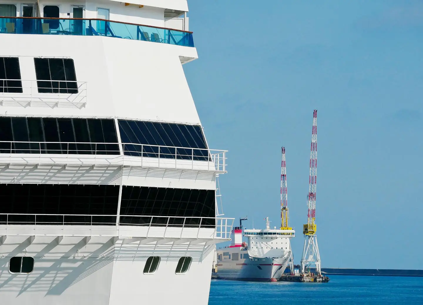 Close-up of a white cruise ship with a port and cranes in the background under a clear blue sky.