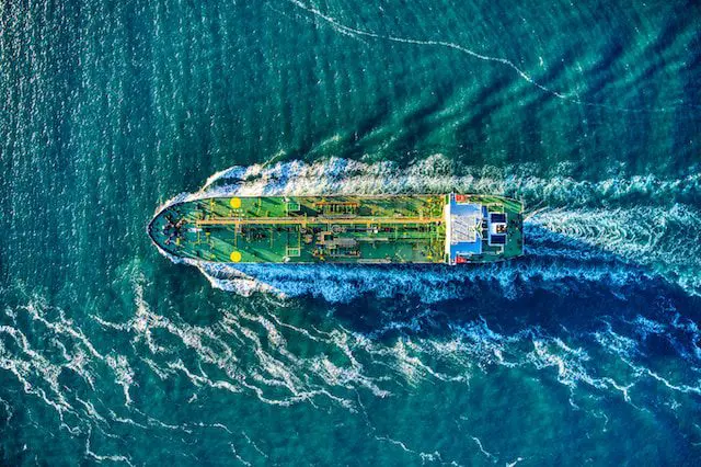 Aerial view of a large cargo ship sailing through blue-green ocean water, leaving waves behind.
