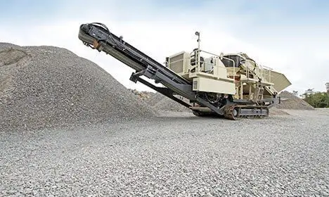 A mobile rock crusher on gravel ground next to large piles of crushed stone under a cloudy sky.