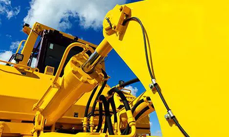 Close-up of yellow hydraulic machinery parts and black hoses against a blue sky with clouds.