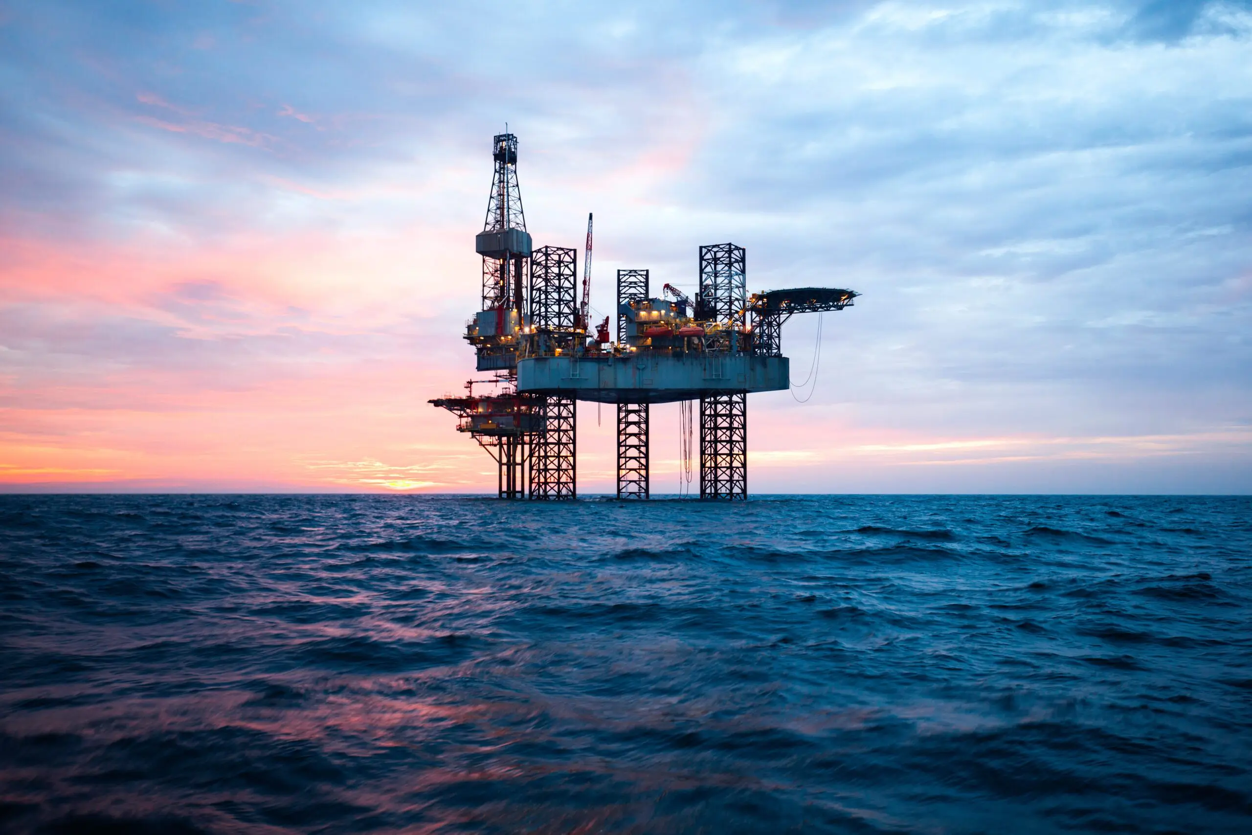 An offshore oil rig stands in the ocean at sunset, with waves in the foreground and a colorful sky.