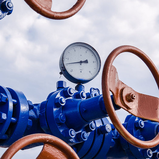 Close-up of a pressure gauge on a blue industrial pipe with brown valve wheels against a cloudy sky.
