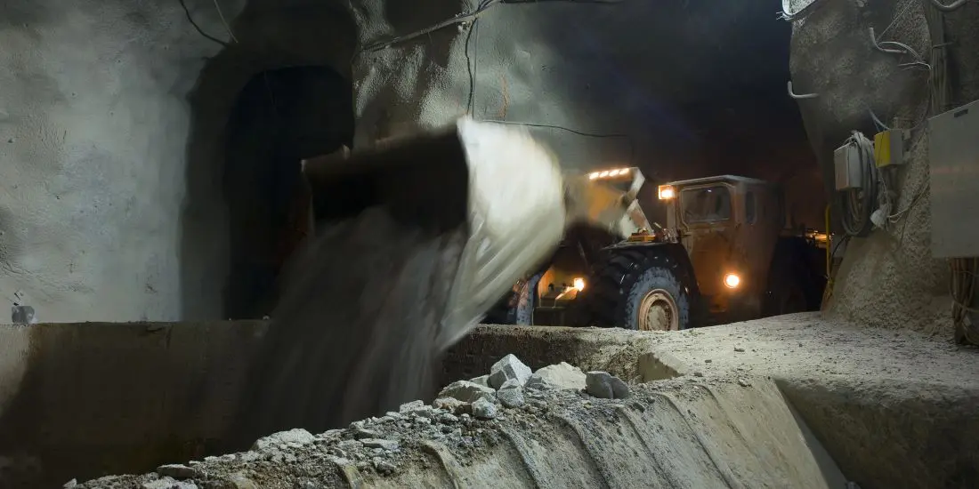 Underground mining vehicle dumping soil into a large container in a dimly lit tunnel.