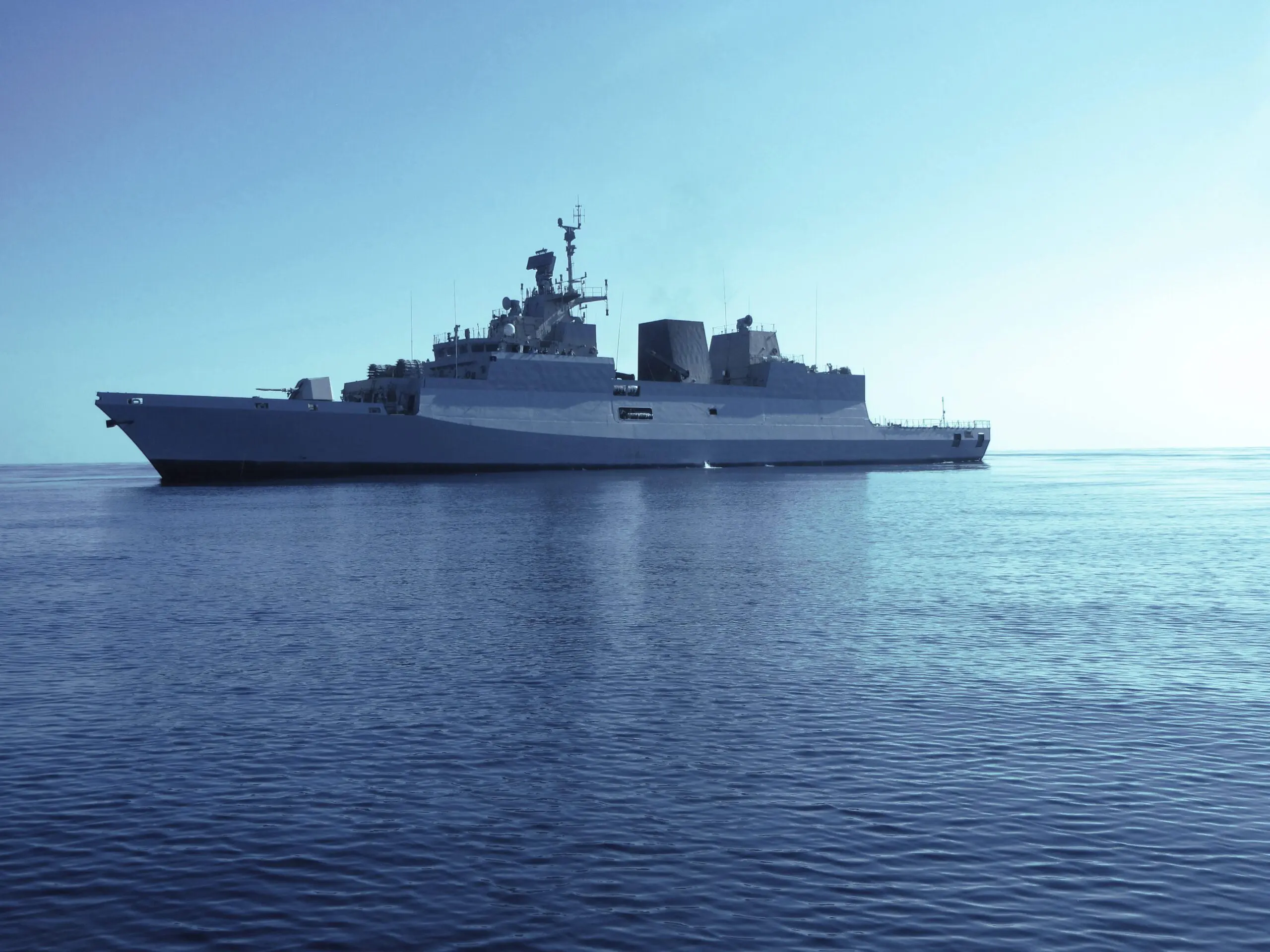 A large navy warship sails on calm blue sea under a clear sky.