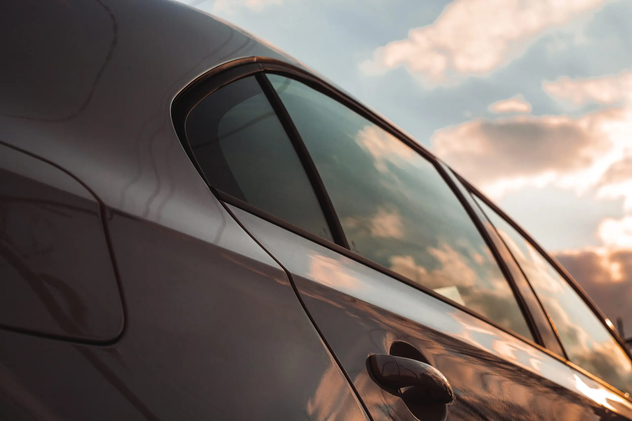 Close-up of a car’s rear window and door reflecting a cloudy sky at sunset.