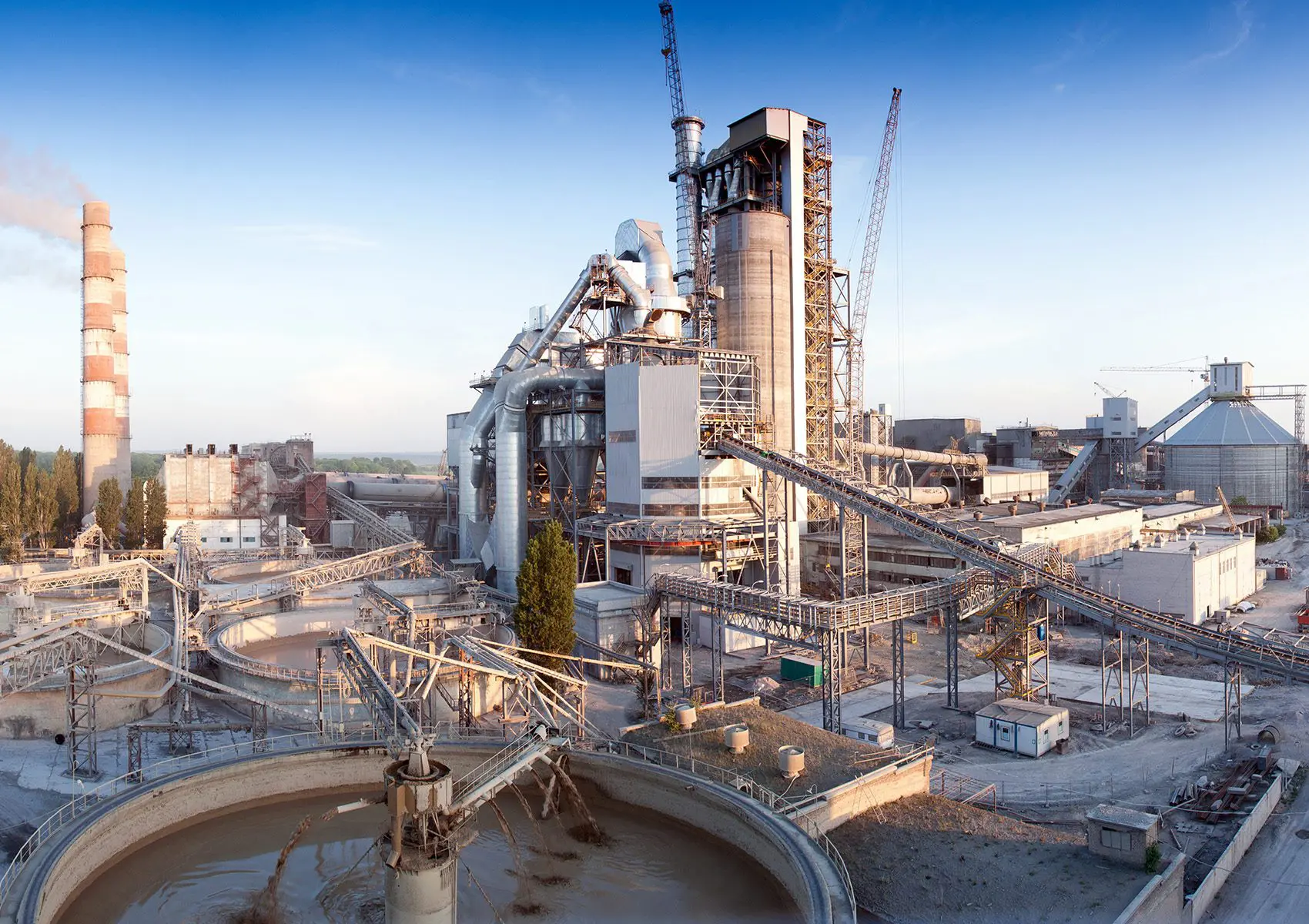 A large industrial cement plant with smokestacks, conveyor belts, and storage silos under a blue sky.