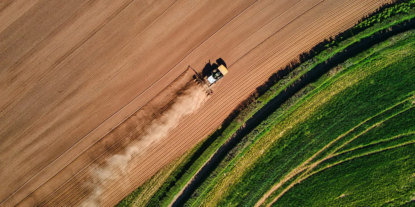 A tractor plows a brown field next to green grass, viewed from above, with dust trailing behind.