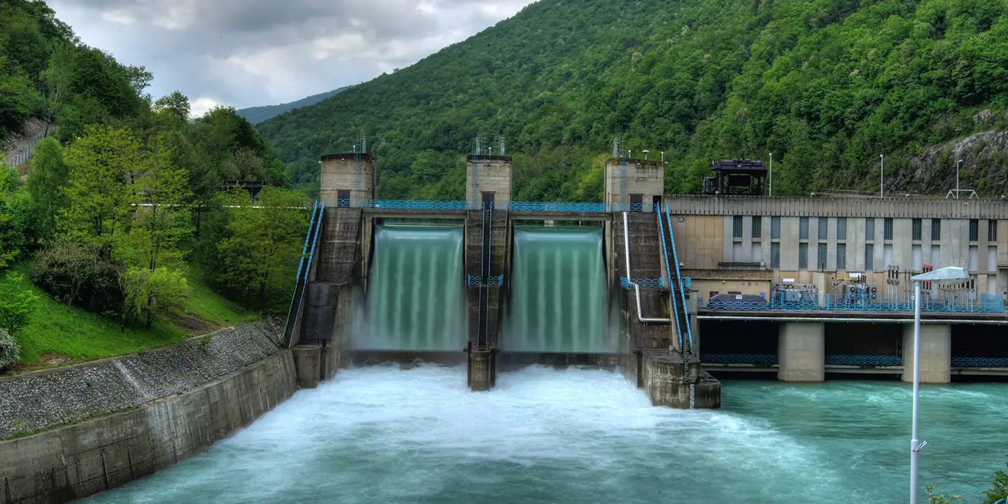 A hydroelectric dam releases water, surrounded by green forested hills and cloudy skies.