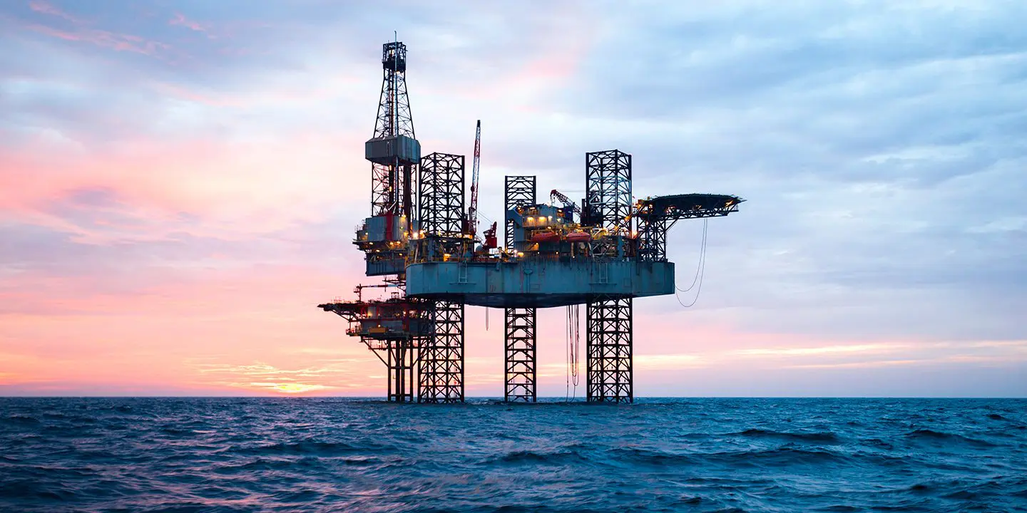 Offshore oil rig stands in the ocean at sunset, with colorful clouds in the sky above.