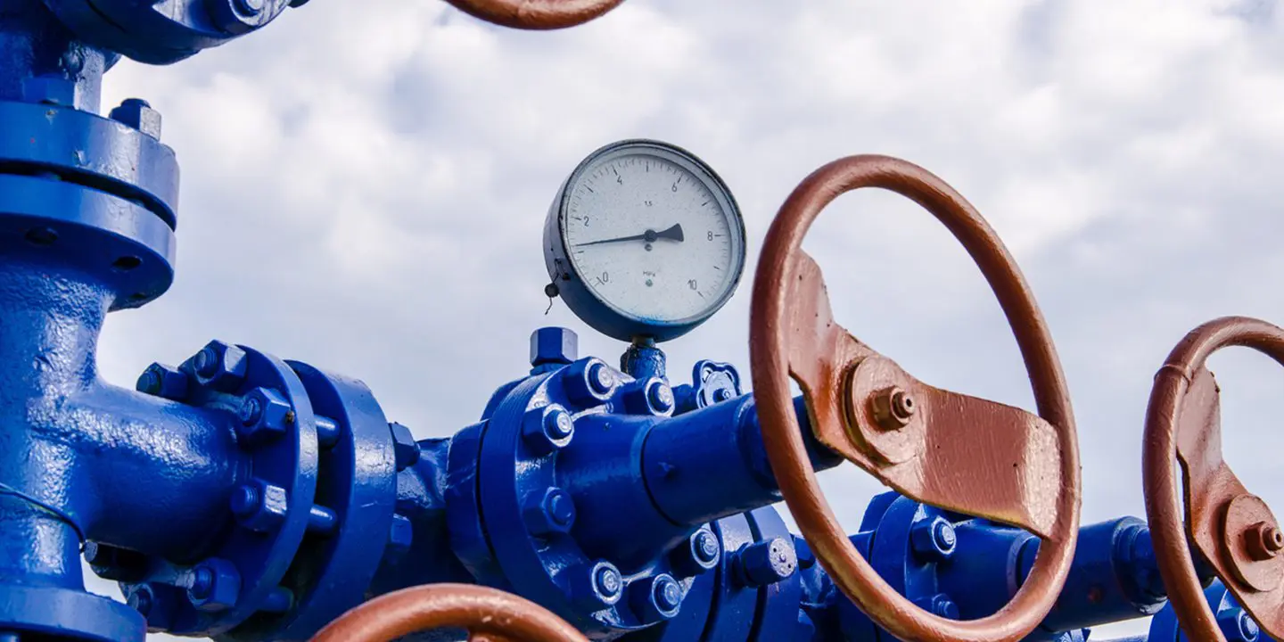 Close-up of blue industrial pipeline with two rust-colored valve wheels and a pressure gauge against a cloudy sky.