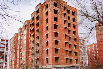 Unfinished red brick apartment building with empty window openings and bare trees in front.