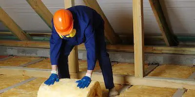 Worker in protective gear installing fiberglass insulation in an attic space with wooden beams.