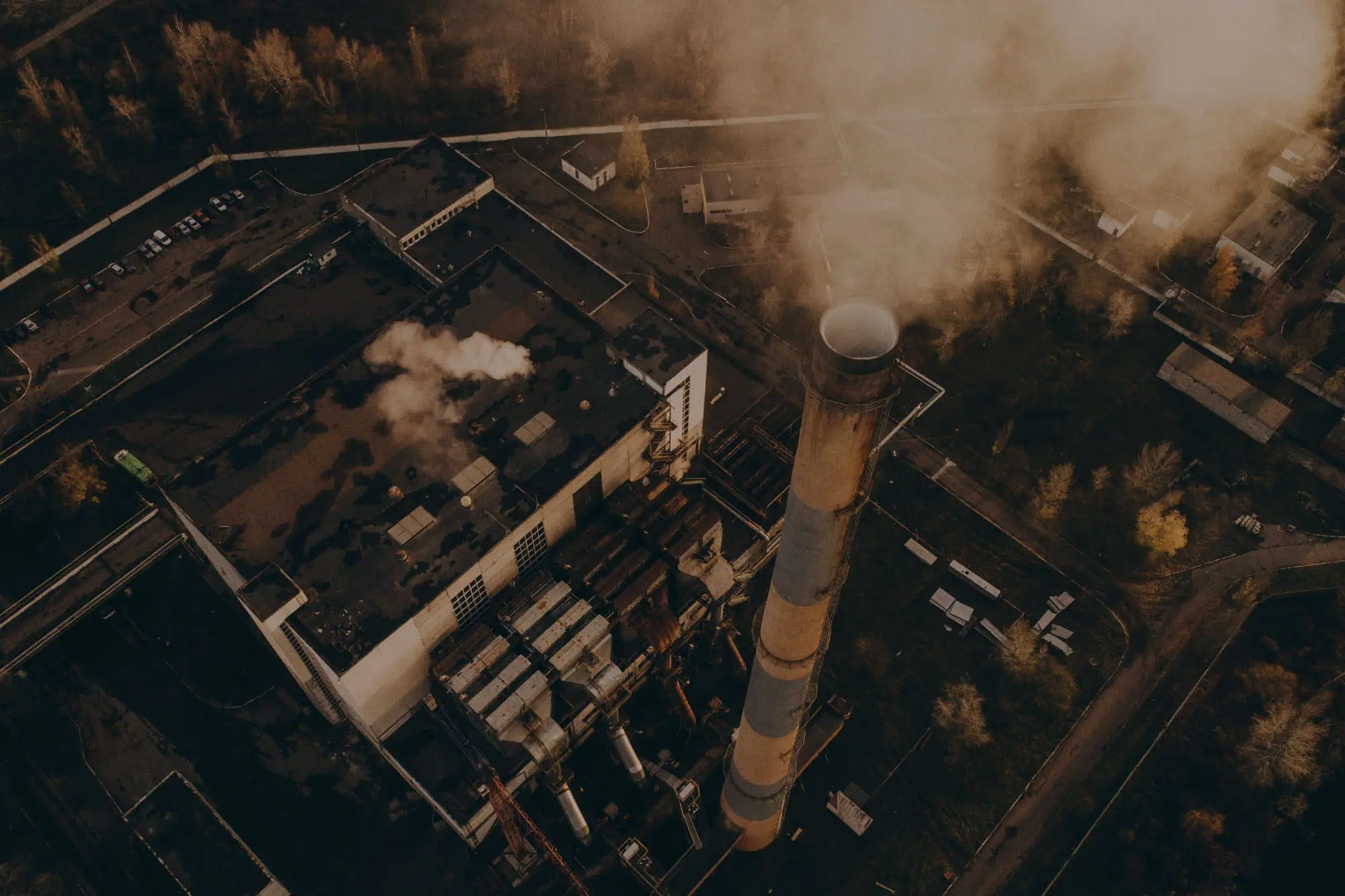 Aerial view of a factory with smoke billowing from a tall chimney, surrounded by trees and buildings.