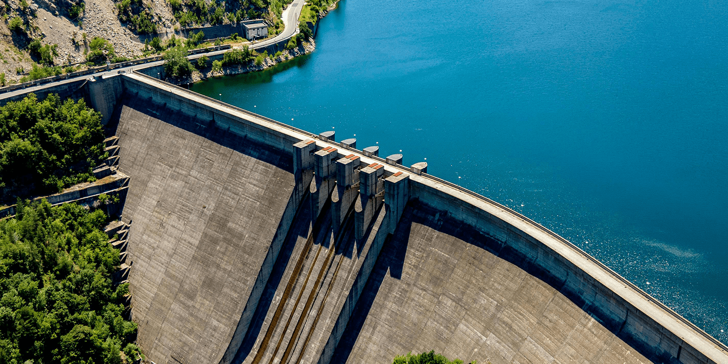 Aerial view of a large concrete dam holding back a blue reservoir, surrounded by greenery.
