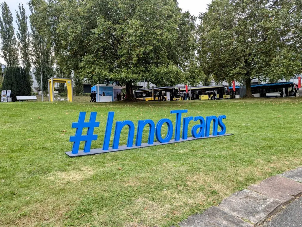 Large blue #InnoTrans sign on grass, with trees, buses, and exhibition booths in the background.