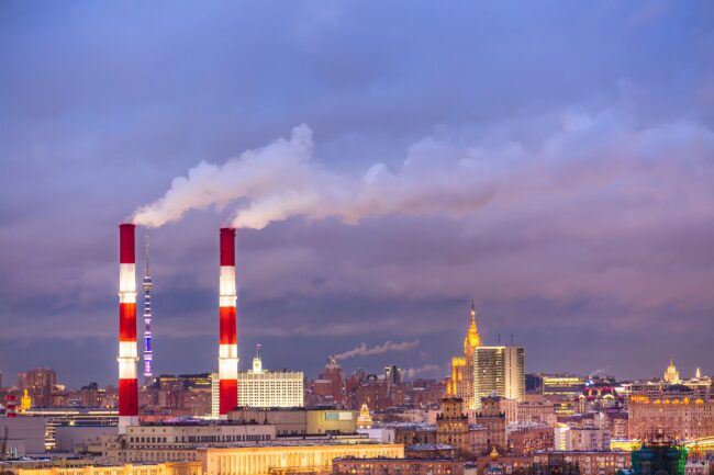 Industrial urban cityscape. Factory pipes of a city landscape in Moscow, Russia during sunset. Two red and white smokestacks emit smoke over a cityscape with various buildings under a cloudy sky at dusk.