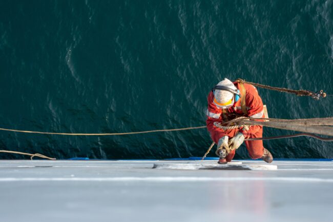 Seaman,Ship,Crew,Working,Aloft,At,Height,Derusting,And,Getting A worker in orange gear climbs a rope on the side of a ship above deep water.