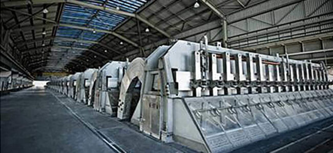 TENMAT-Increases-Portroom-Efficiency-1 Large industrial machinery inside a spacious, high-ceilinged factory building with skylights.