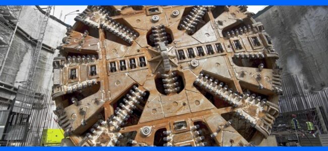 swedens-longest-tunnel-completed-using-ferobide-tenmat A large tunnel boring machine head at a construction site, surrounded by workers and equipment.