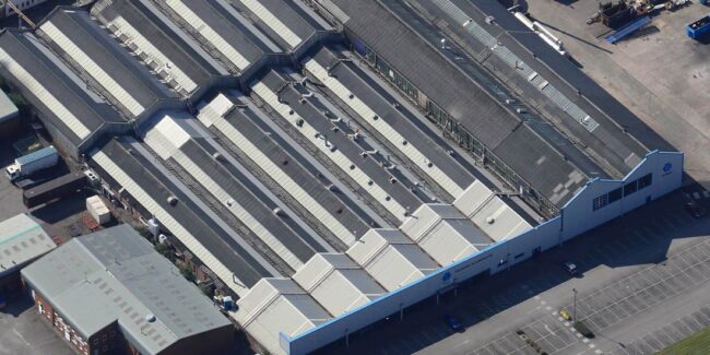 tenmat-hq-manchester1 Aerial view of a large industrial warehouse with a zigzag roof and adjacent parking lot.