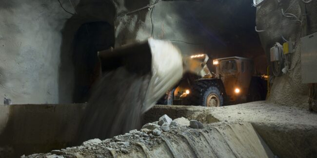 mining-slider Underground mining vehicle dumping soil into a large container in a dimly lit tunnel.