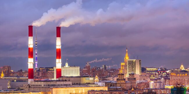 HT-MM-Sectors_Hot-Gas-Filters Two red-and-white smokestacks emit smoke over a cityscape at sunset with buildings in the background.