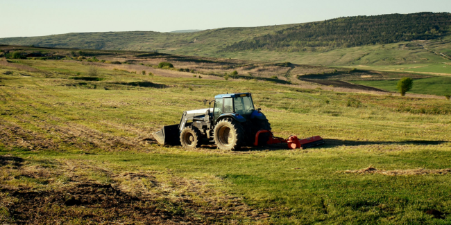 CPM-MM-Materials-Ferobide.jpg A blue tractor plows a grassy field with rolling hills and trees in the background on a sunny day.