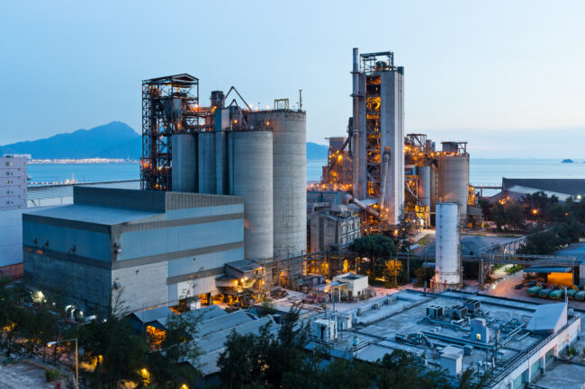 A large industrial factory complex with tall silos and lit structures near water at dusk.