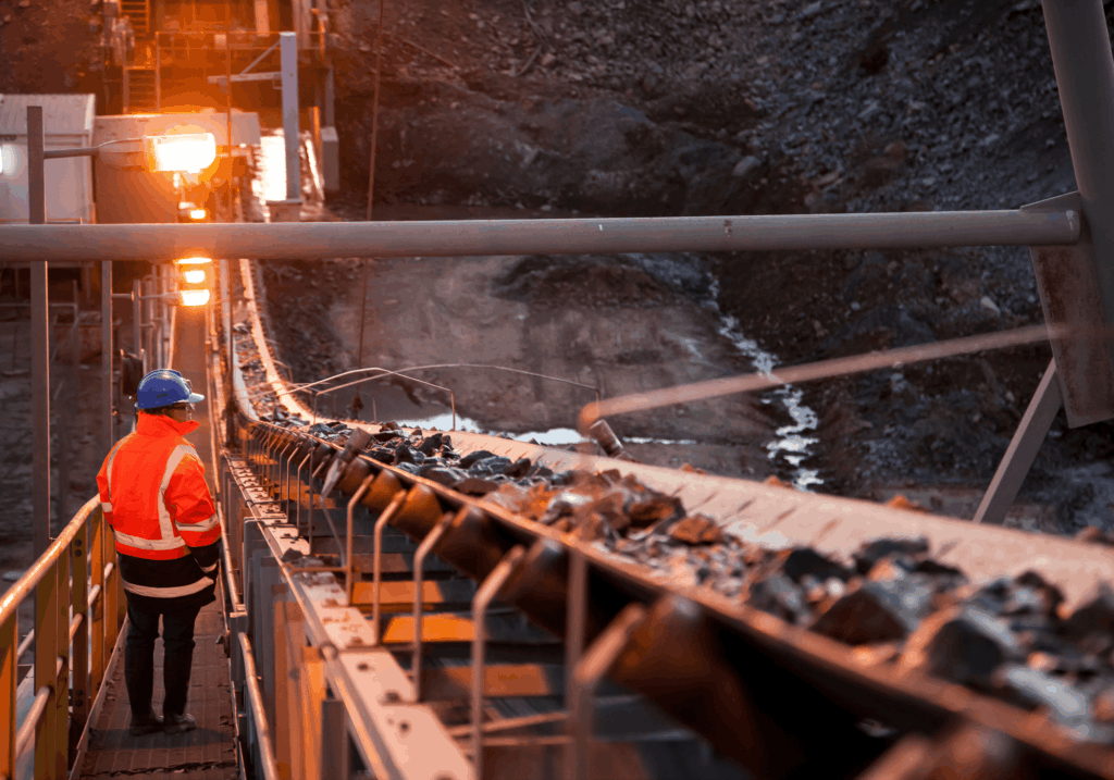 Worker in orange jacket inspects conveyor belt carrying rocks at an industrial mining site.