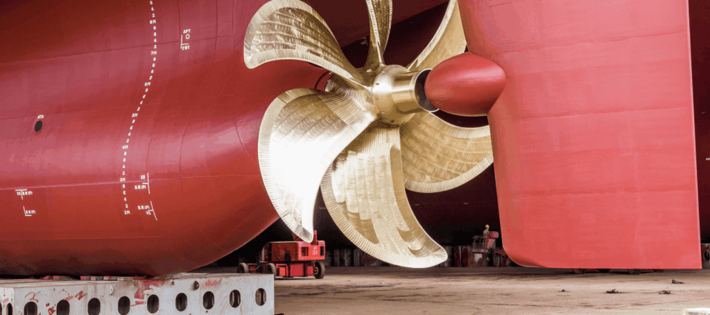 Large gold ship propeller and rudder under a red vessel in a shipyard, with workers nearby.